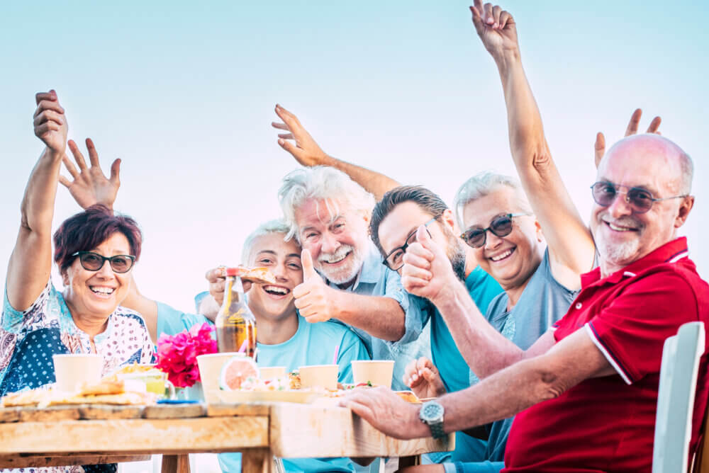 A joyful group of seniors sits at an outdoor table, smiling and raising their hands, enjoying a meal together. - Home Instead