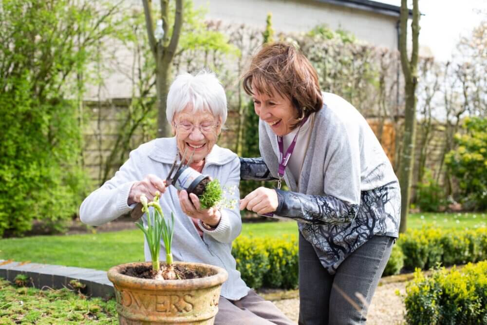 An elderly woman and a younger woman smile while gardening together in a lush, green outdoor setting. - Home Instead