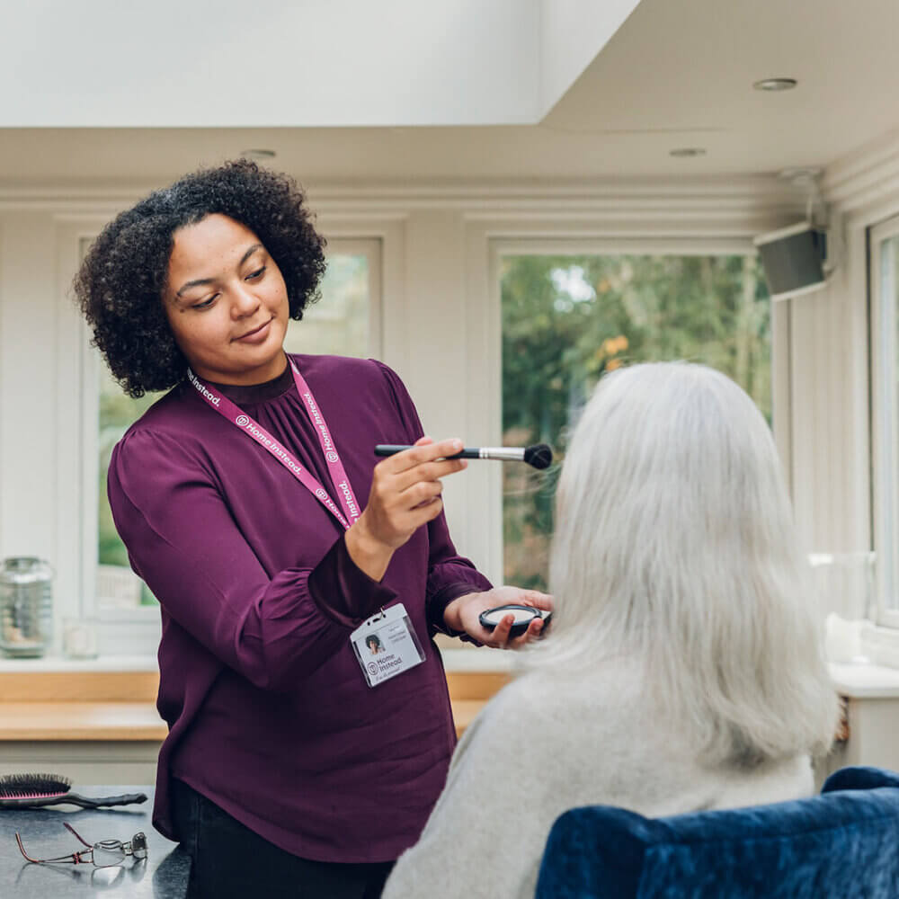 A woman in a purple top applies makeup to an elderly person with gray hair in a brightly lit room. - Home Instead