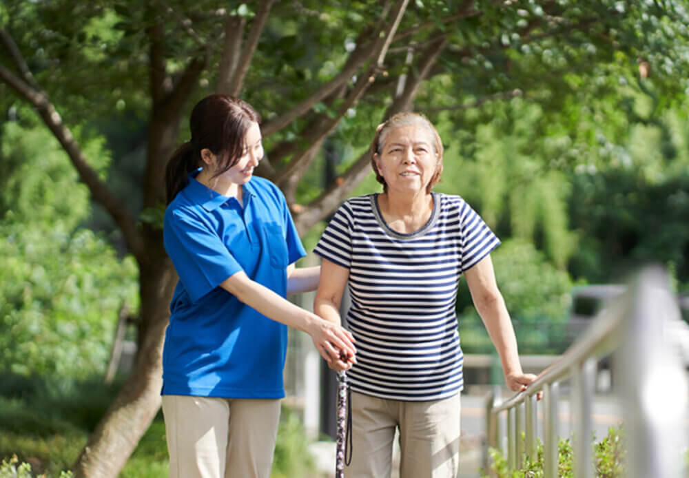 A senior lady holding a walking stick and walking with a care professional.